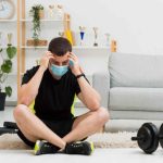 Man wearing gym wear and medical mask sitting next to gym equipment