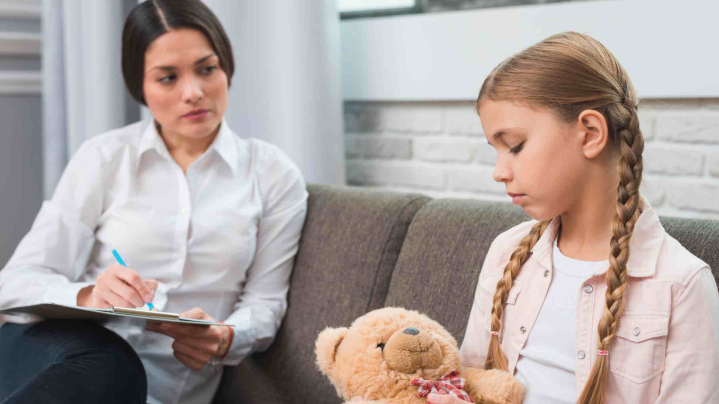 Professional young psychologist observing expressions of her child patient sitting next to her