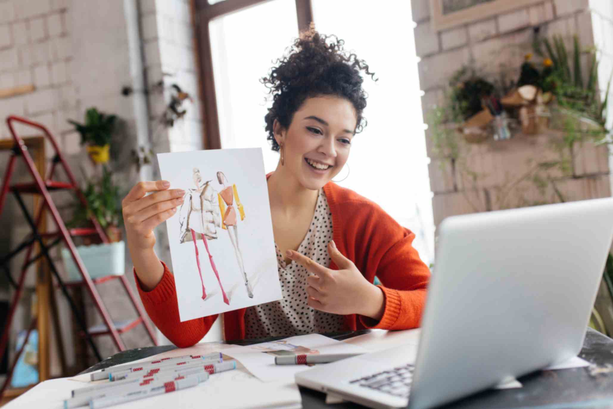 woman with dark curly hair sitting at the table happily showing fashion illustrations in laptop